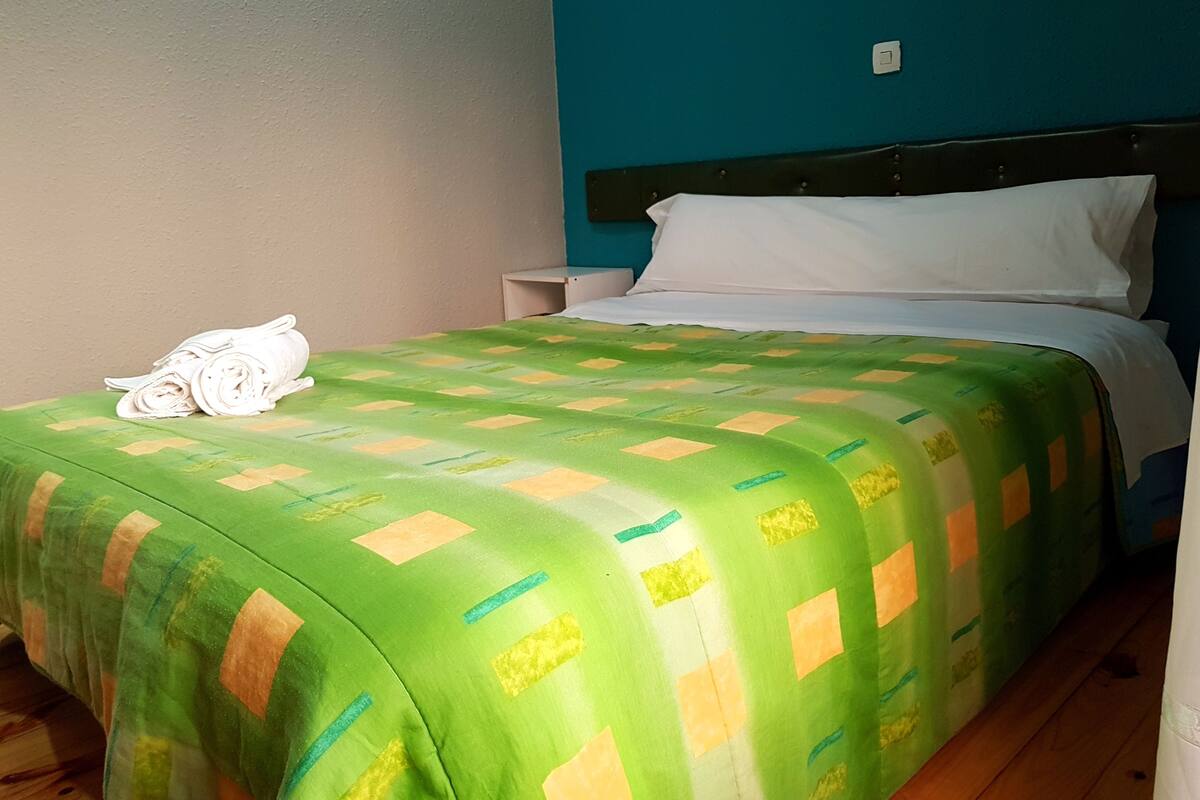 A neatly made bed with a green and yellow patterned bedspread, white pillows, and a stack of white towels on the side.