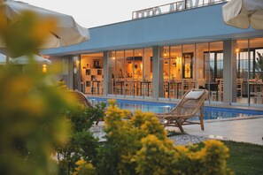 Un edificio moderno con piscina, asientos exteriores y un comedor visible a través de amplios ventanales.
