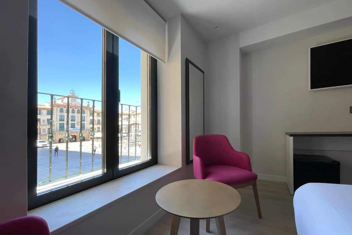 Una habitación de hotel moderna con un ventanal grande, una silla rosa, una mesita redonda pequeña y un televisor de pantalla plana montado en la pared.