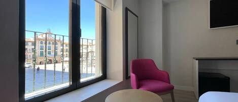 Una habitación de hotel moderna con un ventanal grande, una silla rosa, una mesita redonda pequeña y un televisor de pantalla plana montado en la pared.
