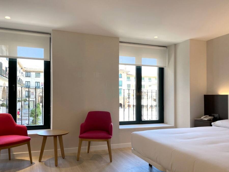 Une chambre d’hôtel avec un grand lit, deux fauteuils rouges, une petite table et une table de chevet avec un téléphone.