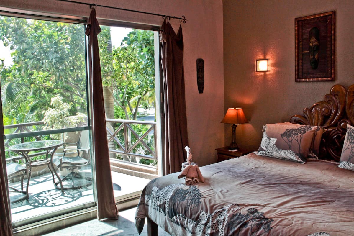 Ein Hotelzimmer mit Balkon, einem Bett mit gemusterten Betttextilien, einem Nachttisch mit Lampe und einem an der Wand hängenden Kunstwerk.