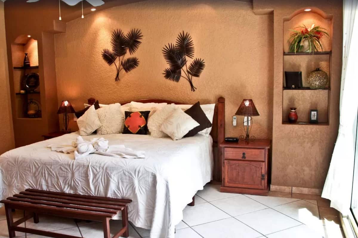 Ein Schlafzimmer mit einem Bett, Nachttischen, einer Bank, Wandbildern und einem Regal mit Dekoration.