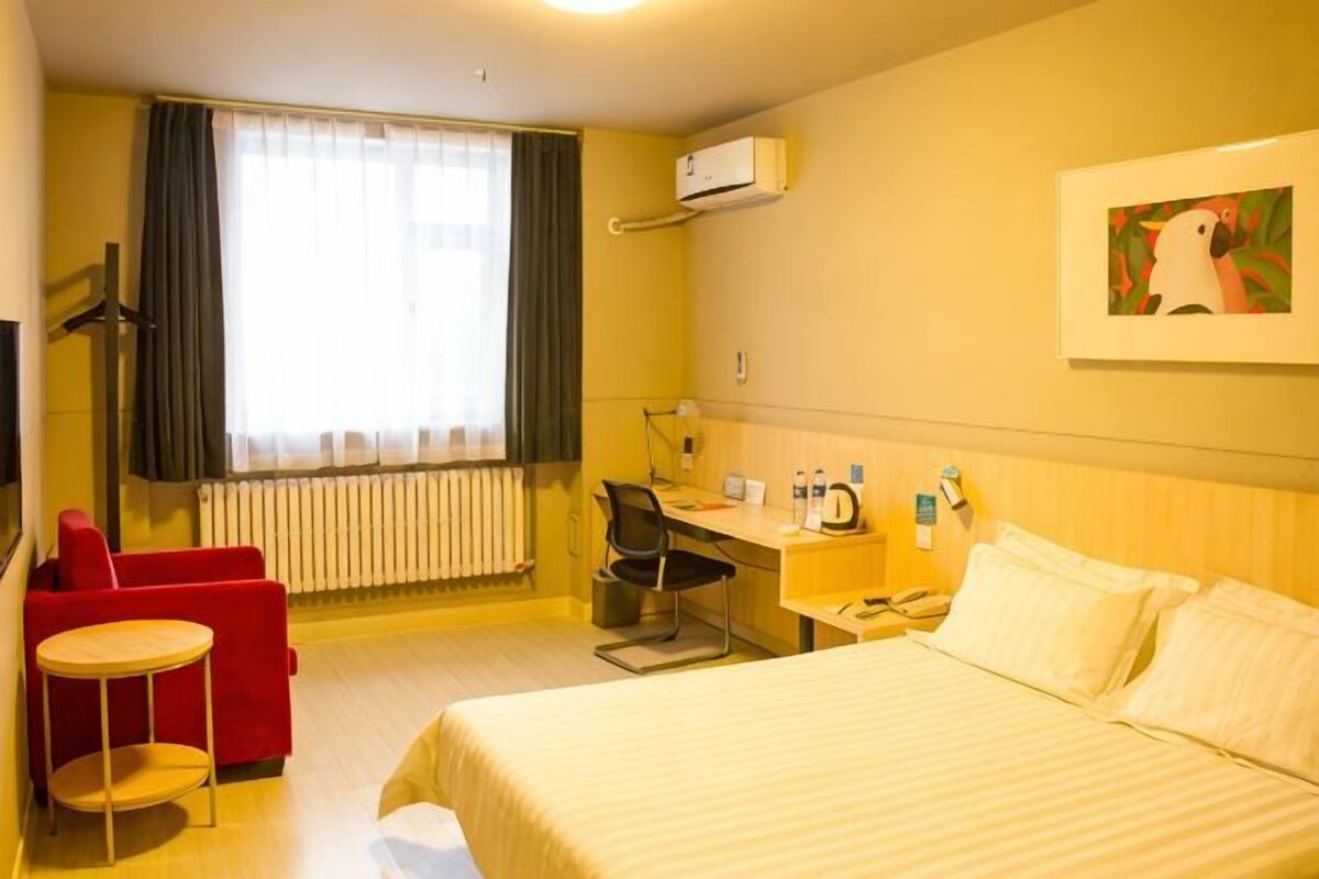 Ein Hotelzimmer mit einem Bett, einem Schreibtisch, einem Stuhl, einem roten Sessel, einem Fenster mit Vorhängen und einem Bild an der Wand.