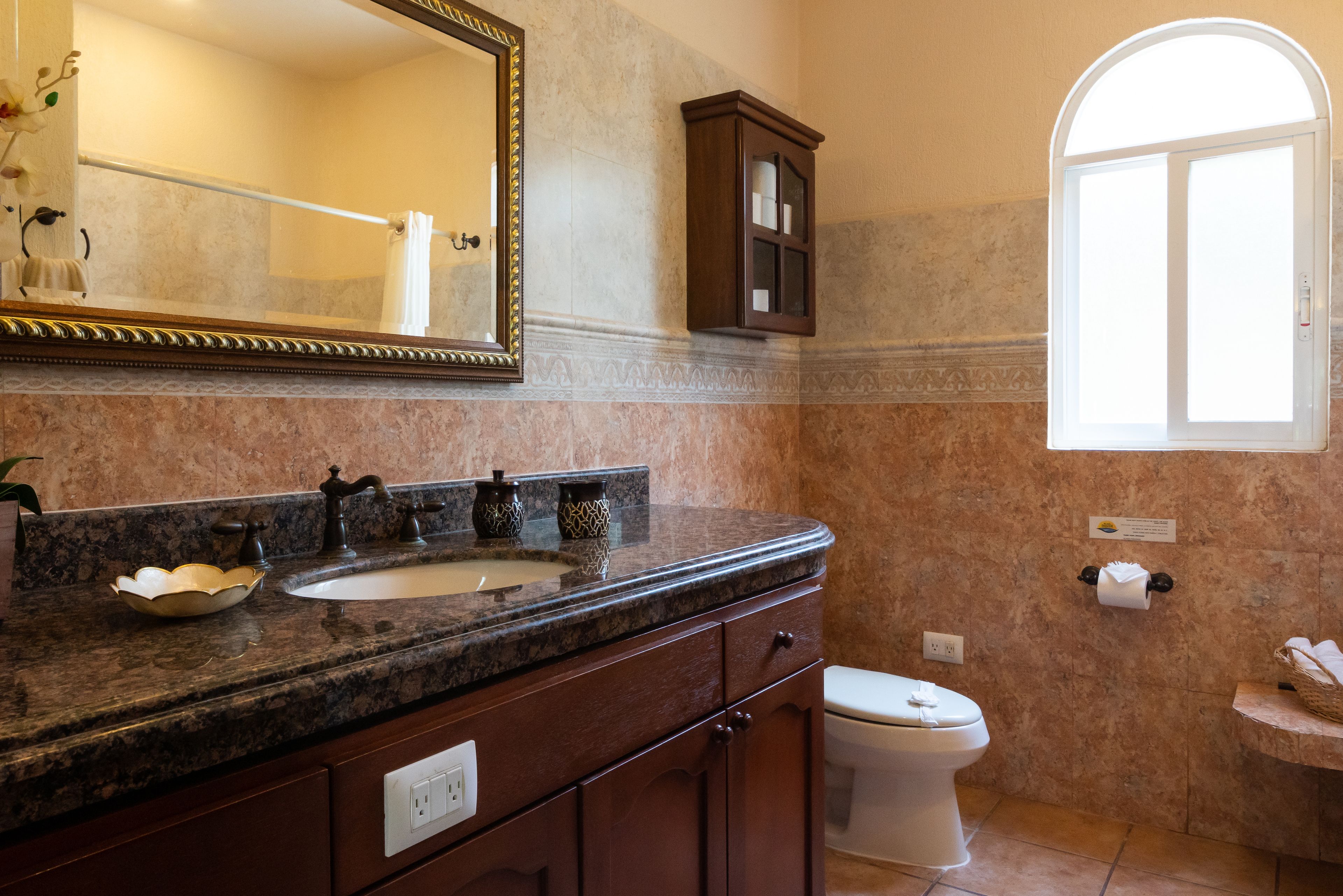 A bathroom with a marble sink, a mirror, a cabinet, a toilet, and a window.