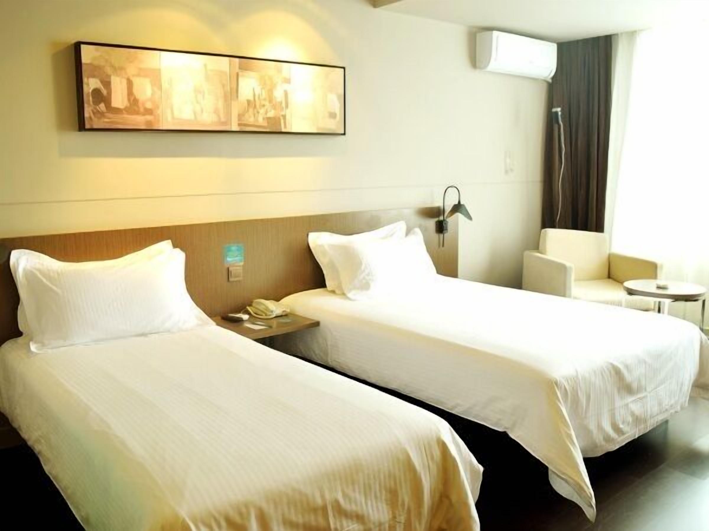 A hotel room with two beds, a small table, a chair, a painting on the wall, and an air conditioning unit.