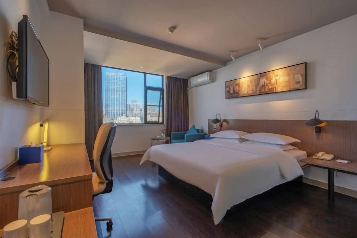 A hotel room with a bed, desk, chair, and a large window with a city view.