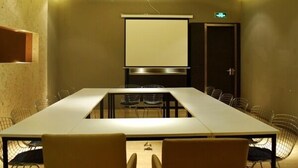 Un salón de conferencias con una larga mesa, sillas, una pantalla de proyector y una puerta con letrero de salida.