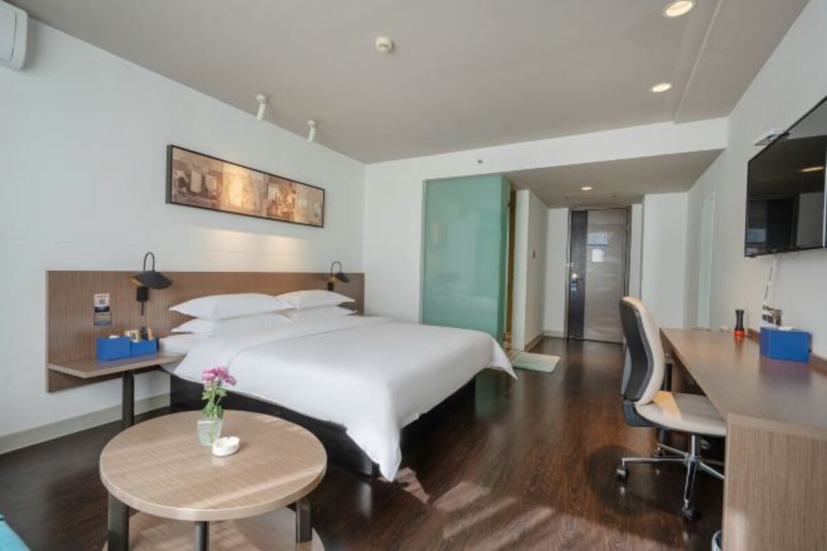 A modern hotel room with a large bed, a desk, a chair, a small round table with a vase of flowers, a painting on the wall, and a television.