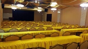 Un salón de conferencias con filas de mesas y sillas, equipado con luces en el techo y una pantalla de proyector.