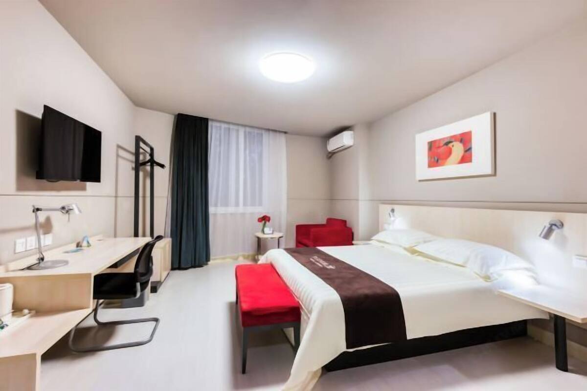 A hotel room with a bed, a desk, a chair, a TV, a window with curtains, and a painting on the wall.