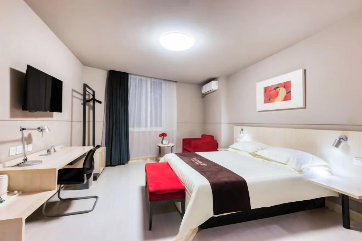 A hotel room with a bed, a desk, a chair, a TV, a window with curtains, and a painting on the wall.