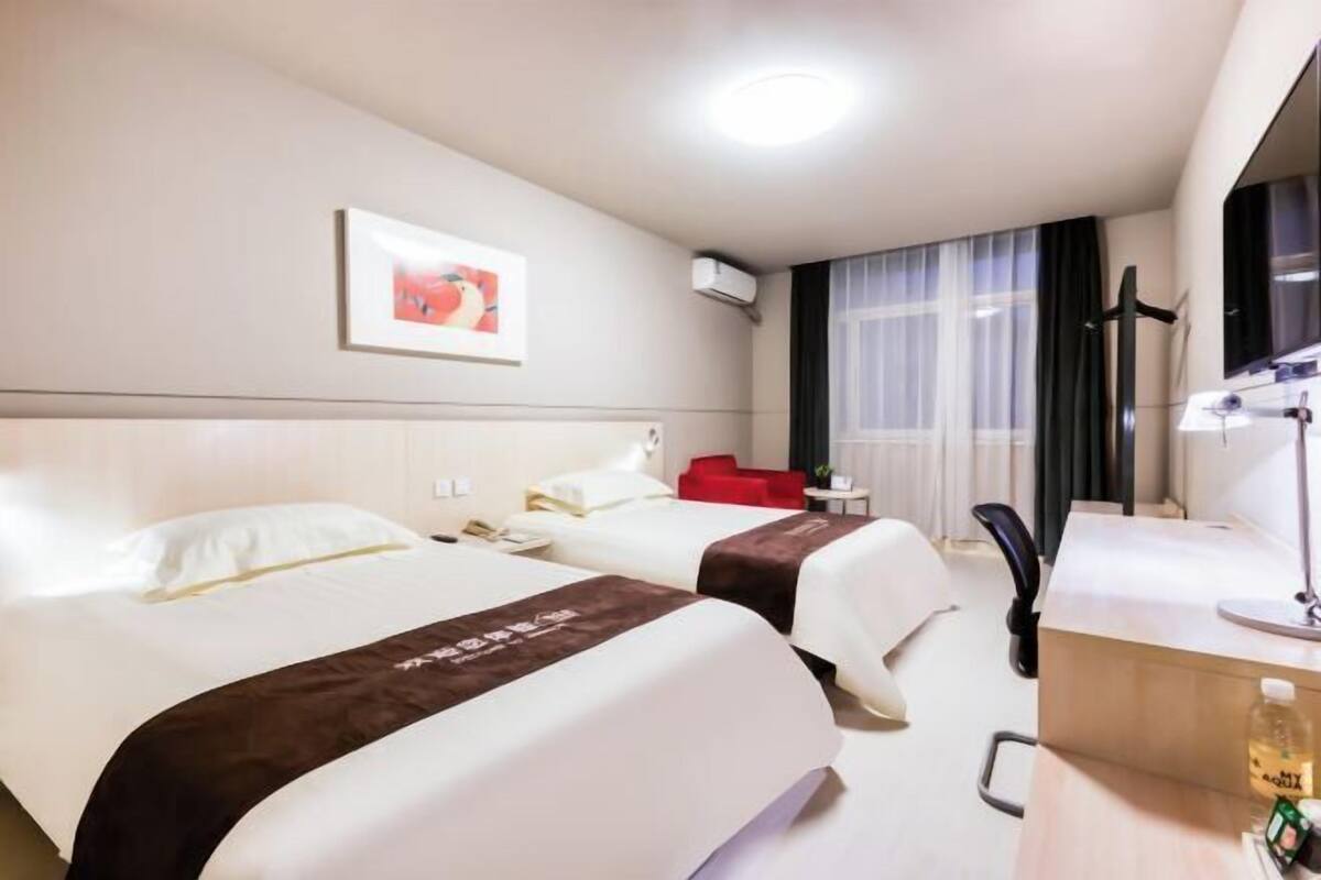 A hotel room with two beds, a desk, a chair, a desk lamp, a bottle of water, and a framed picture on the wall.