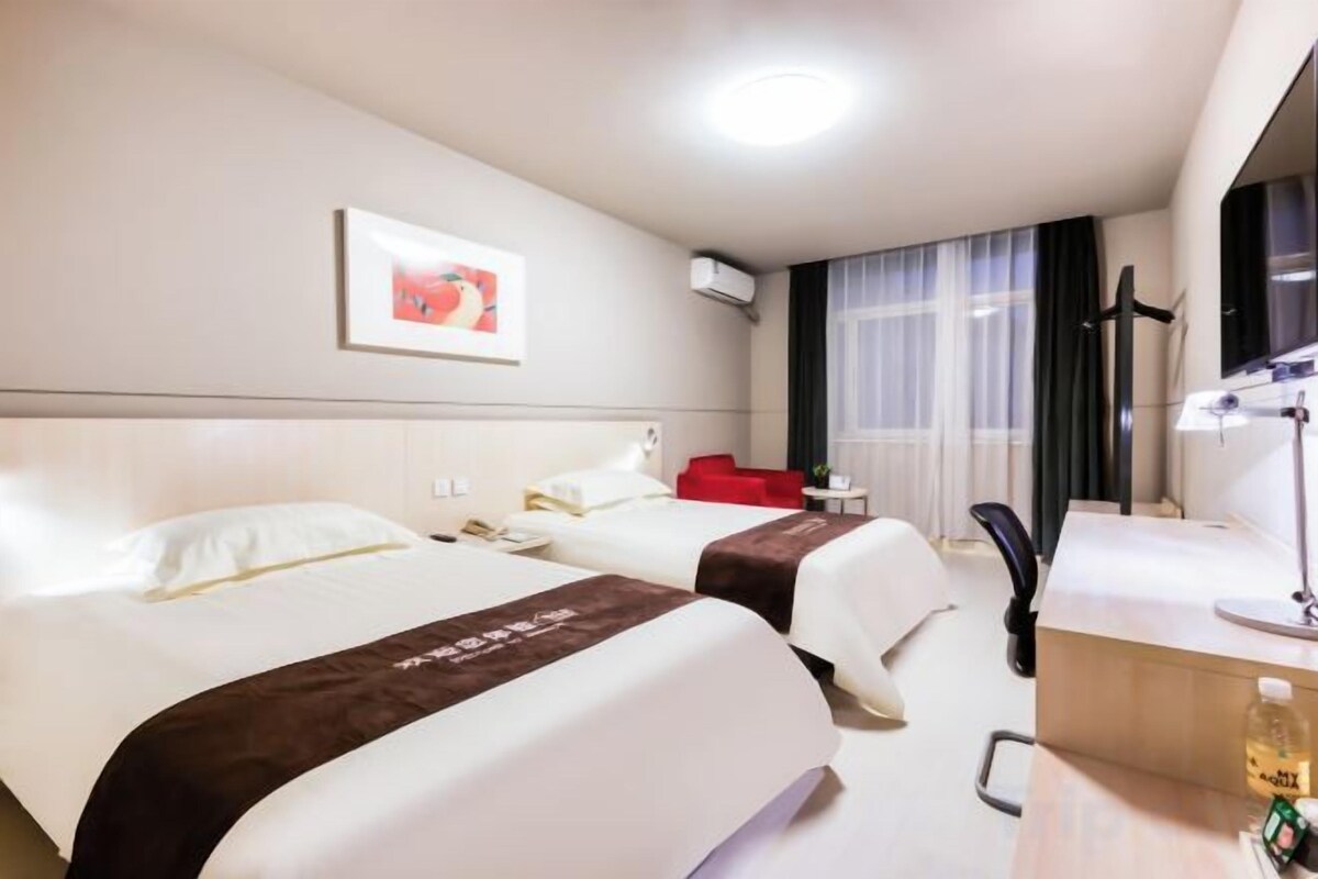A hotel room with two beds, a desk, a chair, a window with curtains, and a wall-mounted air conditioner.