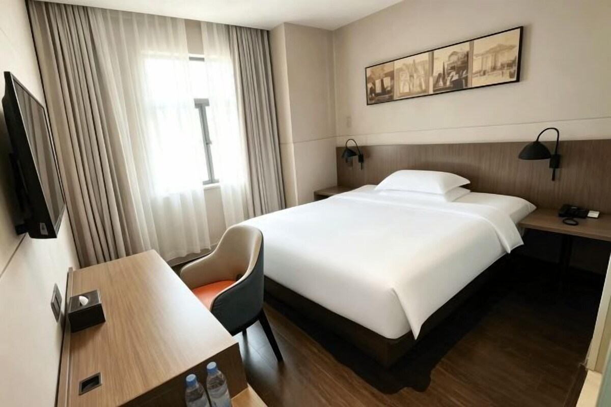 Habitació d'hotel amb un llit, escriptori, cadira, televisió i finestra amb cortines.