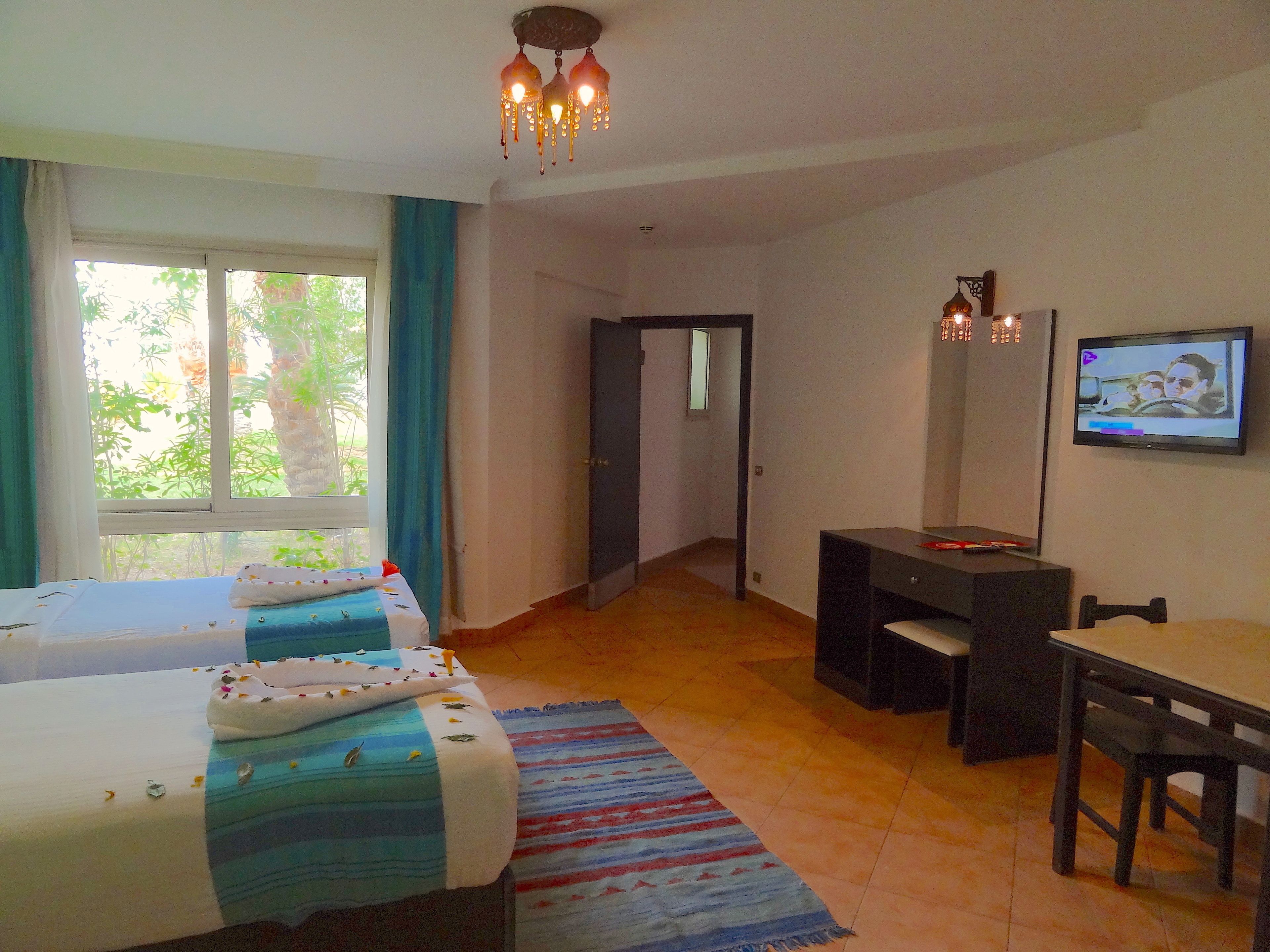 A hotel room with two beds, a desk, a chair, a television, and a view of greenery outside.