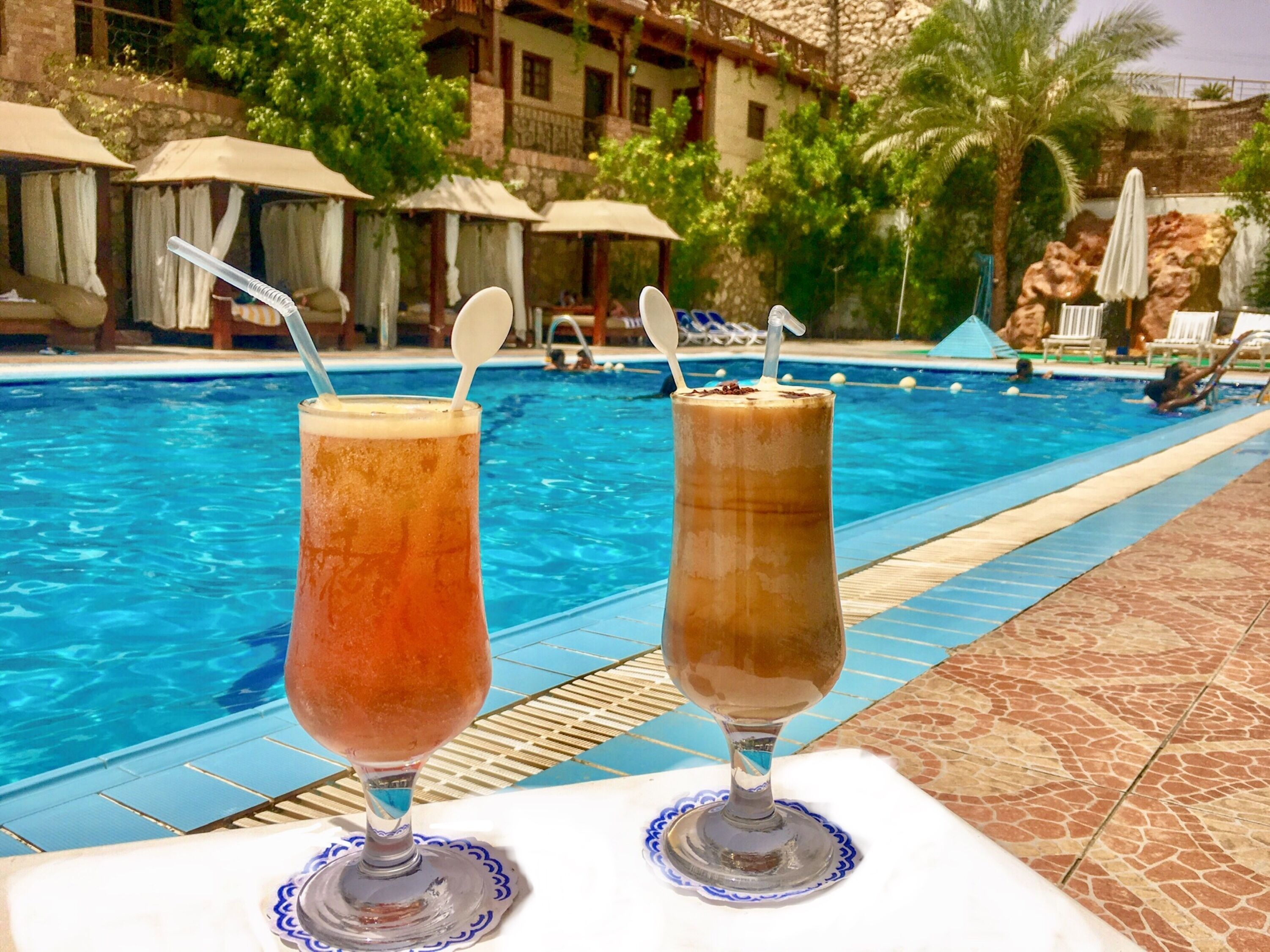 Two drinks on a table by a poolside with umbrellas and lounge chairs in the background.