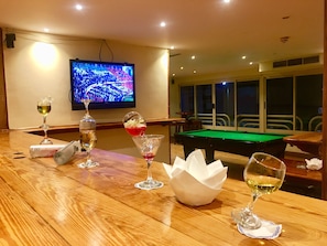 Une table de billard dans une pièce où se trouve un téléviseur diffusant un événement sportif, un bar en bois avec des verres et des bouteilles, et une fenêtre donnant sur l’extérieur.