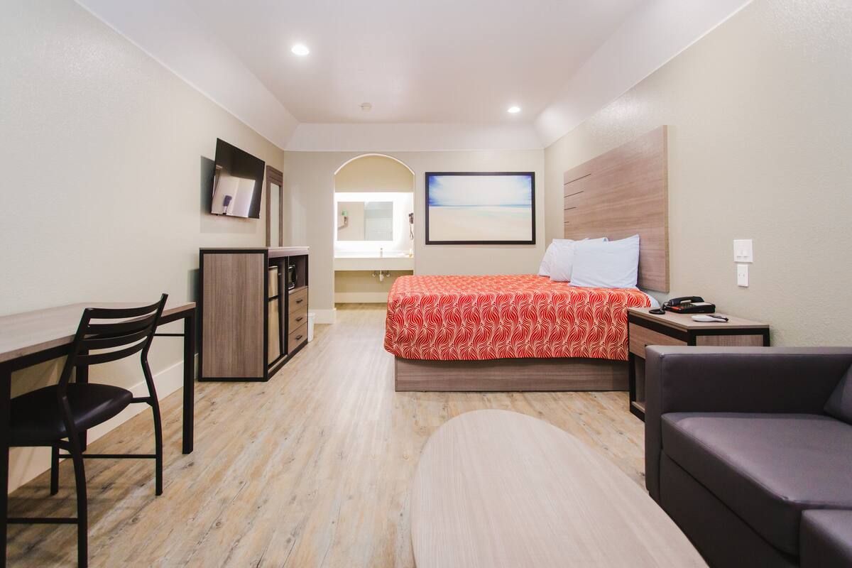 A hotel room with a bed, a desk, a chair, a sofa, a round table, a TV, and a painting on the wall.