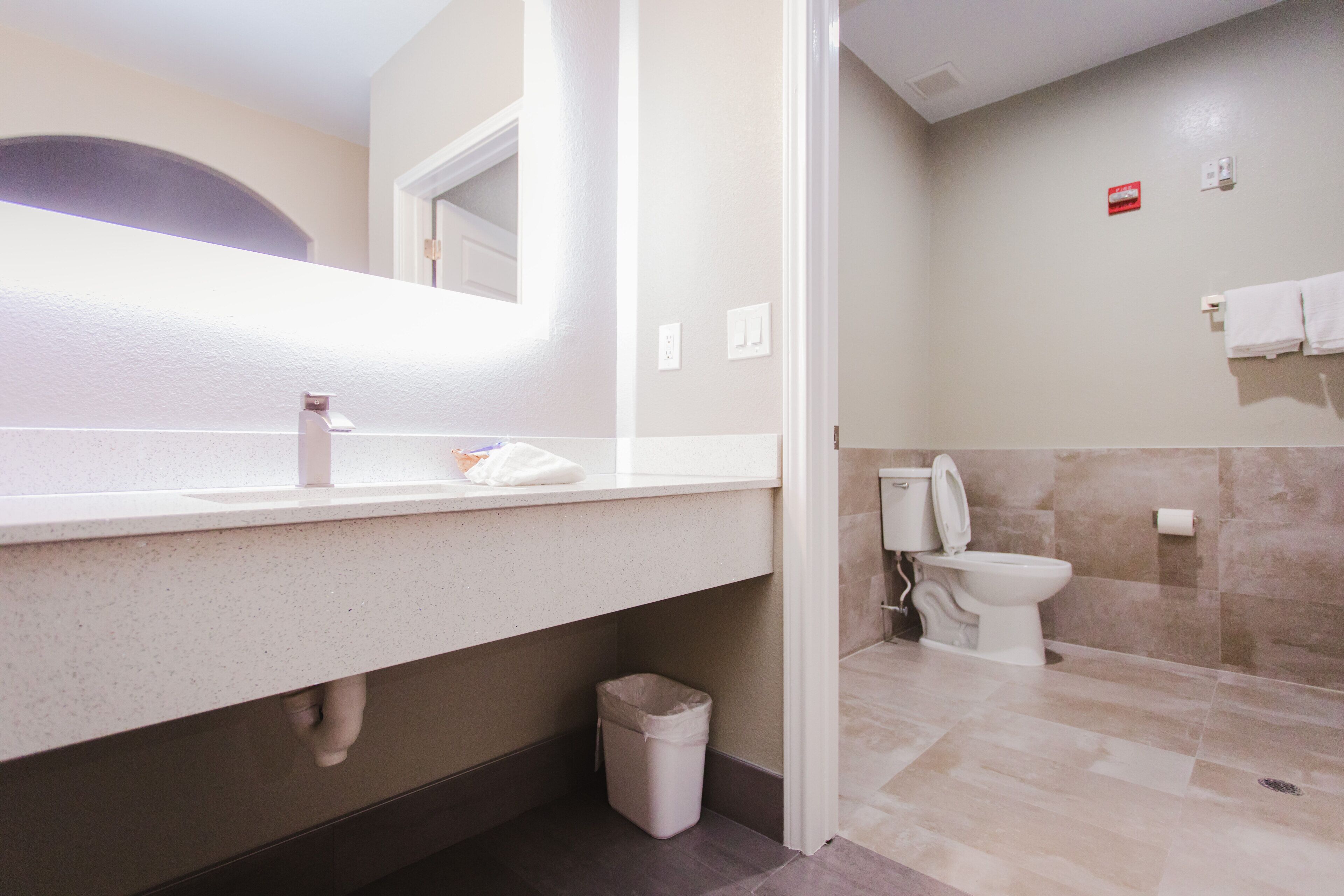 A modern bathroom with a white countertop, a sink, a mirror, a toilet, and a trash bin.