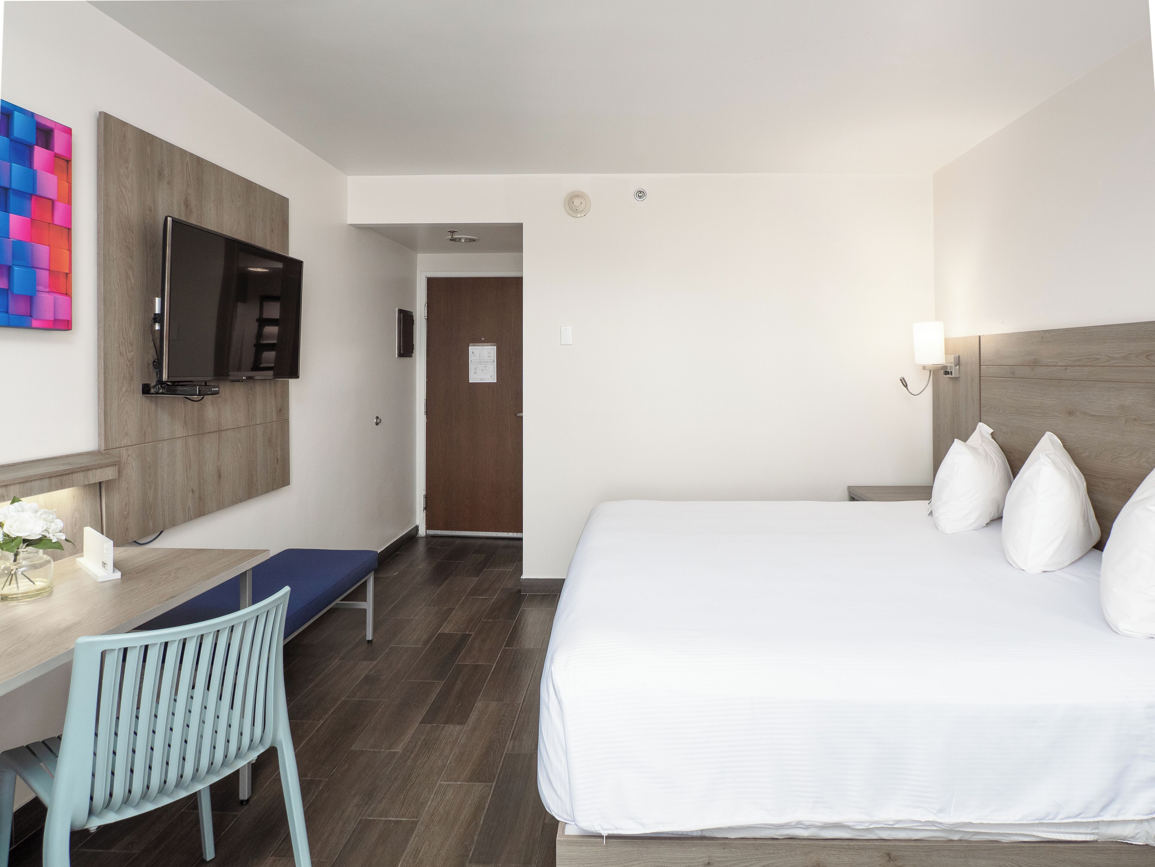 A hotel room with a bed, a desk with a chair, a TV mounted on the wall, and a wooden headboard.