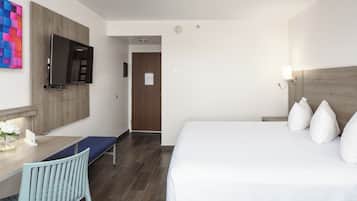 A hotel room with a bed, a desk with a chair, a TV mounted on the wall, and a wooden headboard.