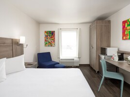 Quarto de hotel com cama, escrivaninha, cadeira, micro-ondas, cafeteira e janela com cortinas.