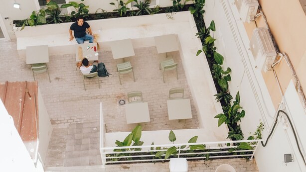 An outdoor patio area with white tables and chairs, potted plants, and a person sitting on a bench. - El Patio de Chueca - Hostel (Madrid)