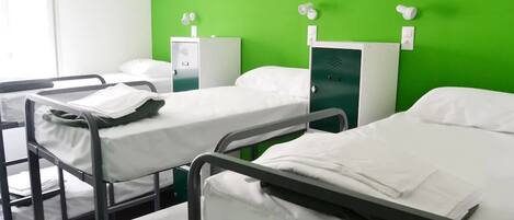 A hospital room with three beds, each with white linens and a black metal frame. There is a green wall with a medical device mounted on it.