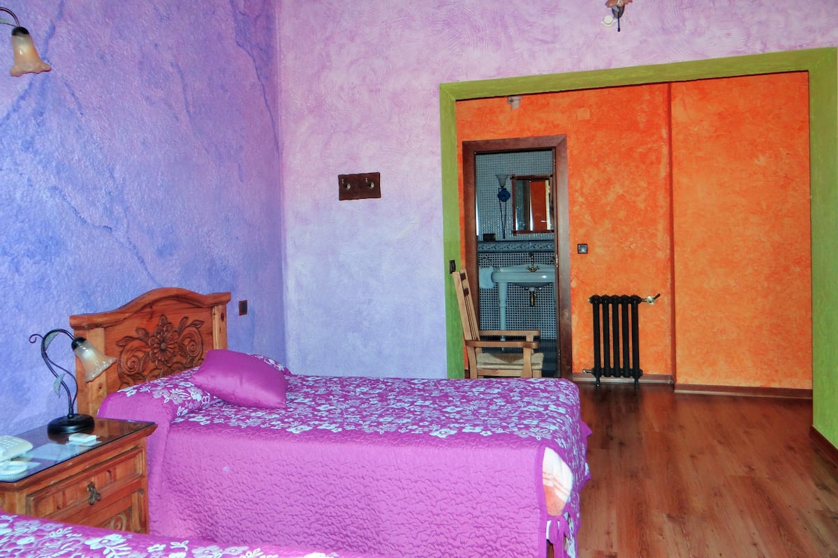 Un dormitorio con cama de madera, mesita de noche, silla, radiador y baño con lavabo y espejo.