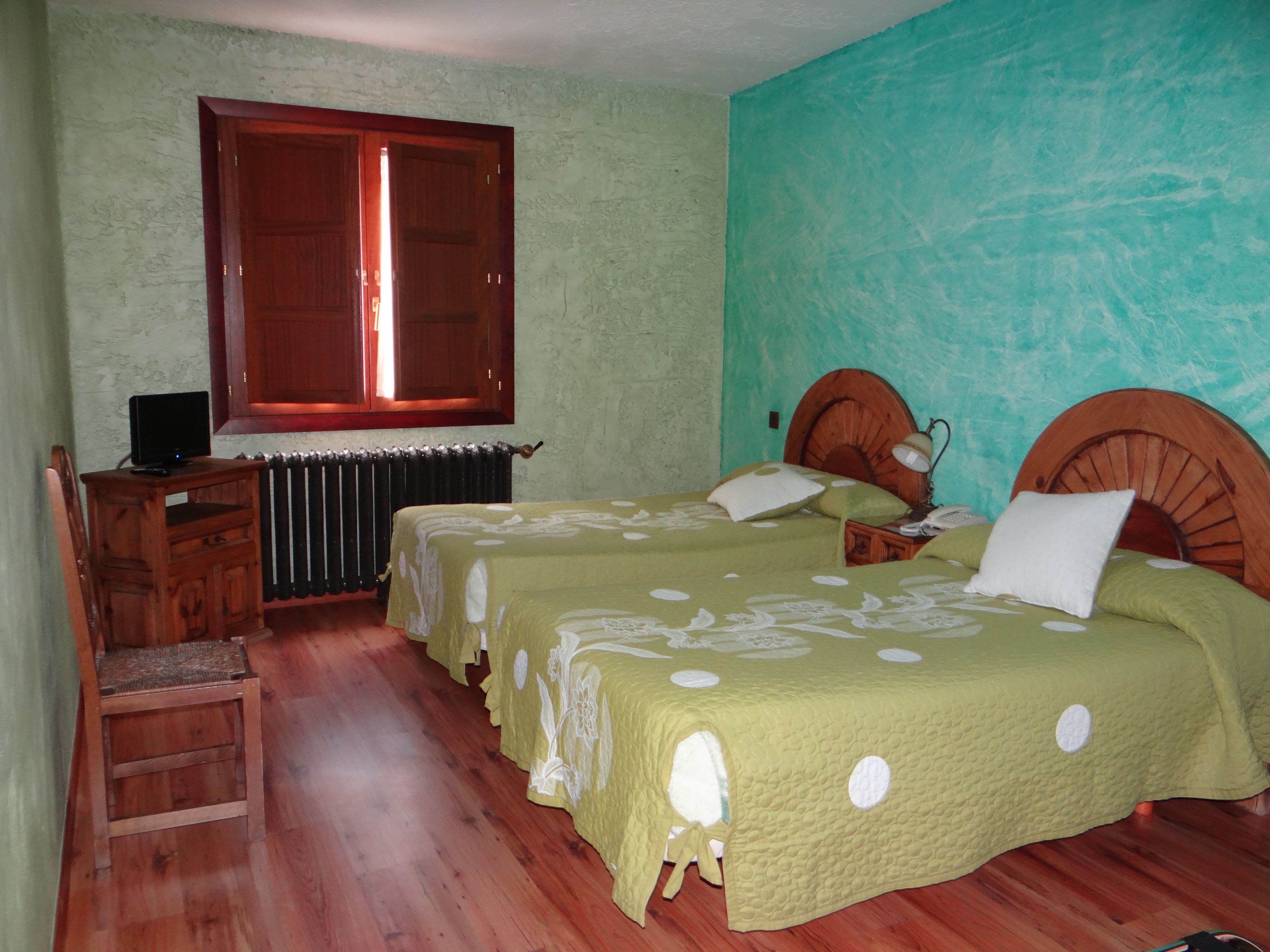 A room with two beds, a wooden desk, a chair, a radiator, and a window with wooden shutters.