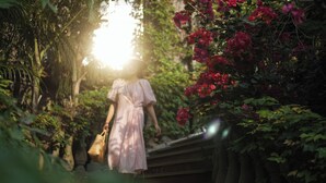 Une femme vêtue d’une robe rose se promène dans un jardin où poussent des fleurs rouges et de la verdure.