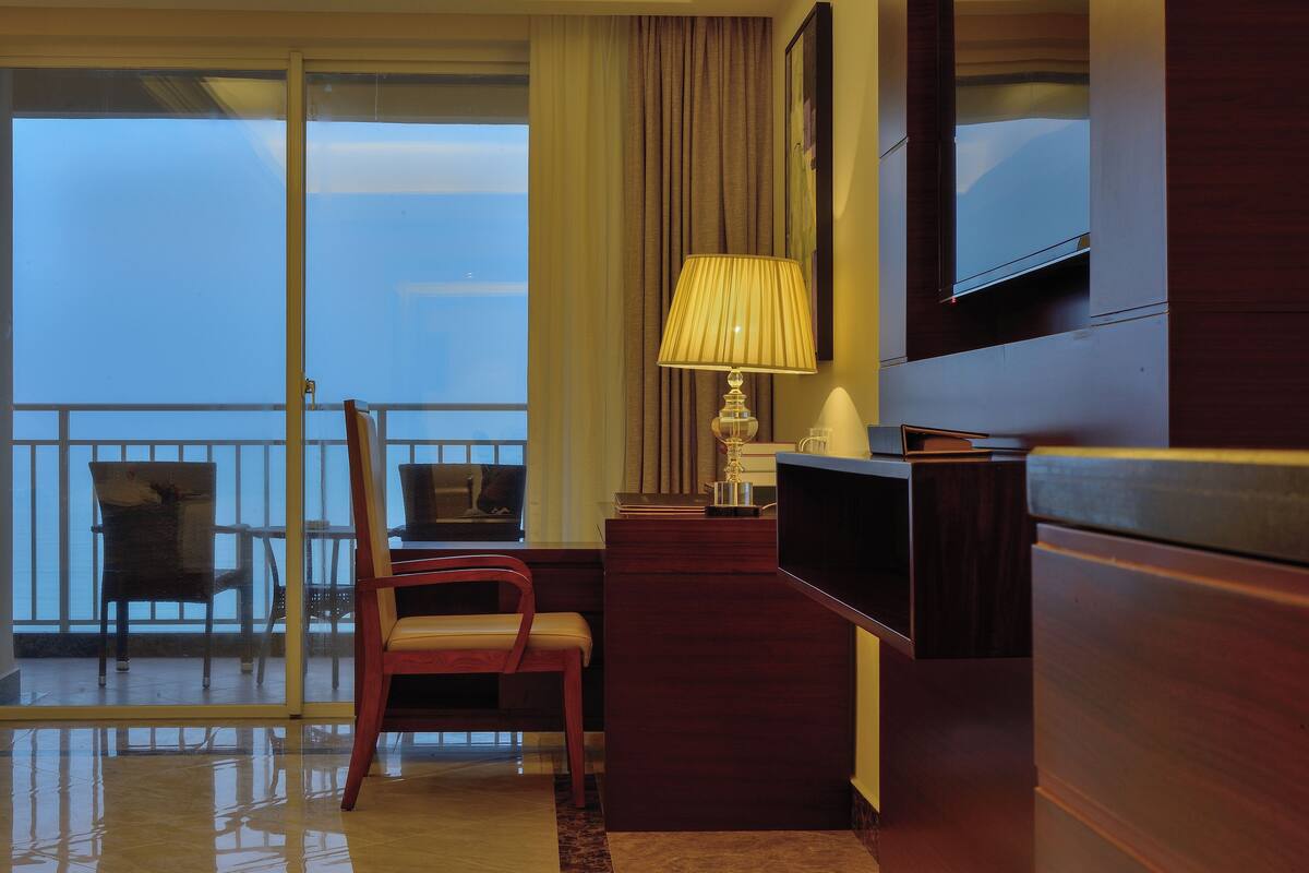 A hotel room with a desk, chair, TV, and a view of the city through large windows.