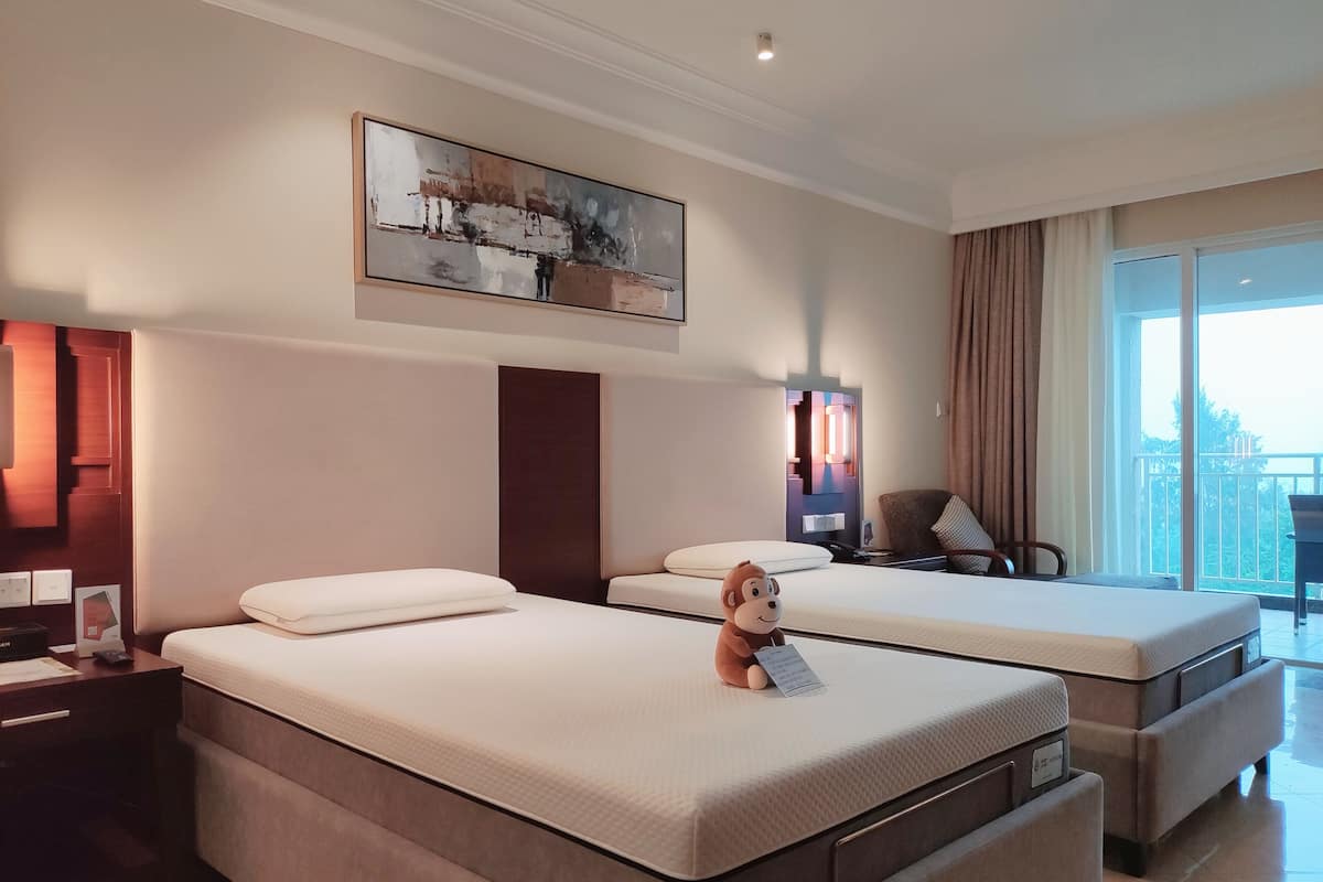 A hotel room with two beds, a desk, a chair, a window with a view, and a painting on the wall.