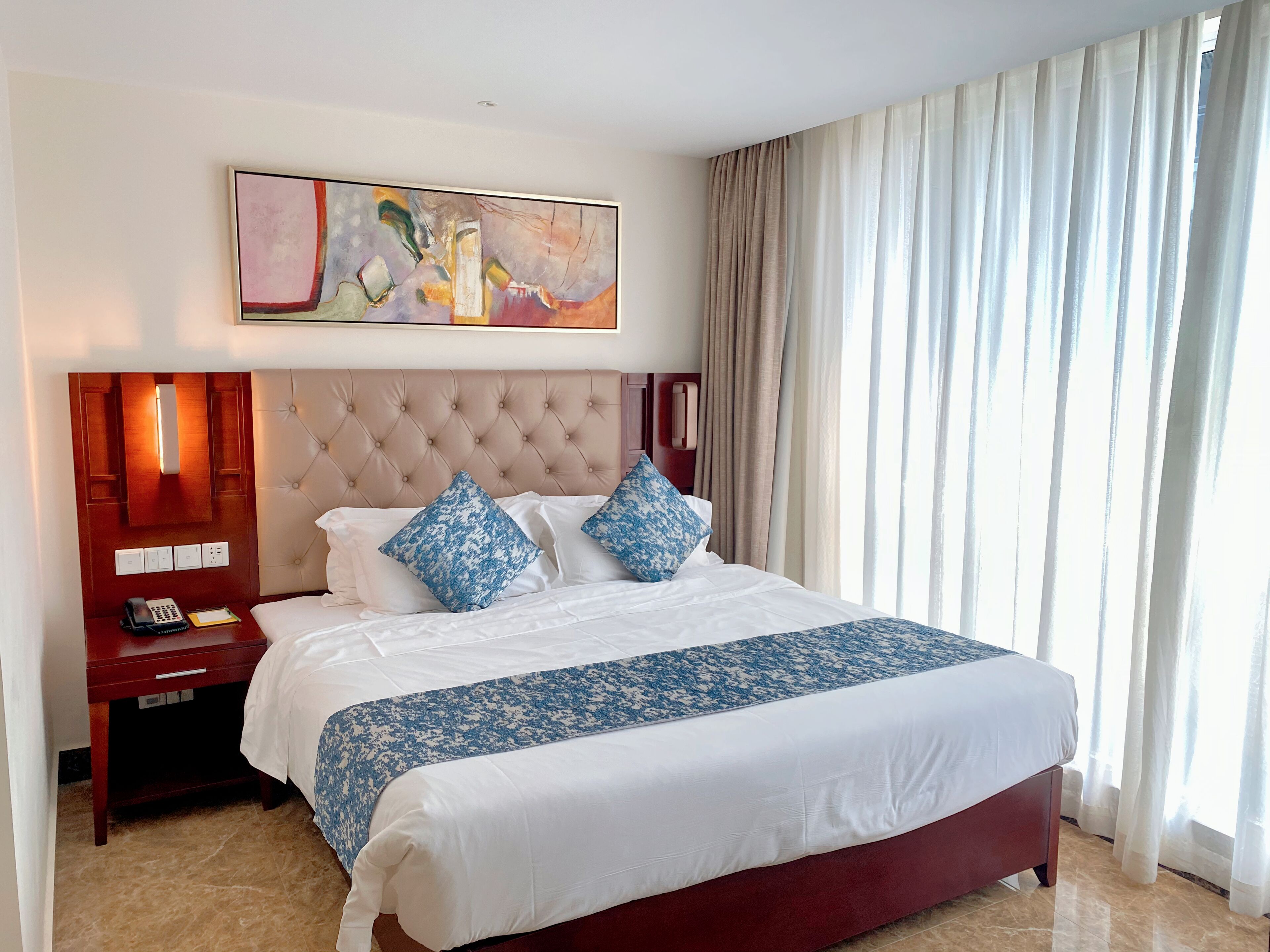 A hotel room with a large bed, a nightstand with a phone, a painting above the bed, and a window with curtains.