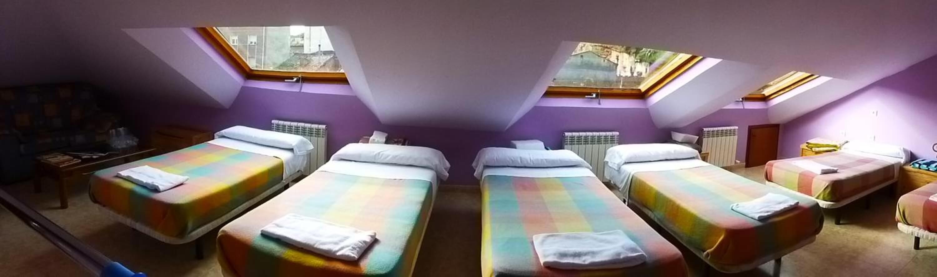 Una habitación con cuatro camas dispuestas en fila, cada una con un edredón de colores. Hay tragaluces sobre las camas que dejan entrar luz natural.