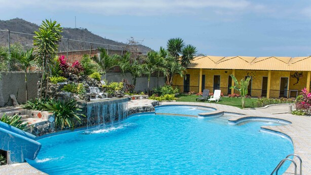 A swimming pool with a waterfall feature, surrounded by tropical plants and a yellow building with multiple doors. - Tuzco Lodge (Puerto Lopez)