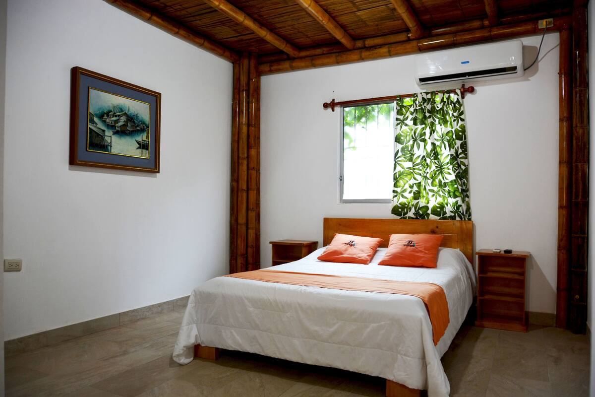 A bedroom with a bed, wooden headboard, two nightstands, a window with curtains, an air conditioning unit, and a framed picture on the wall.