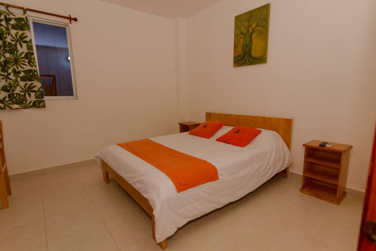 A neatly made bed with an orange blanket, a wooden nightstand with a lamp, and a painting of a tree on the wall.