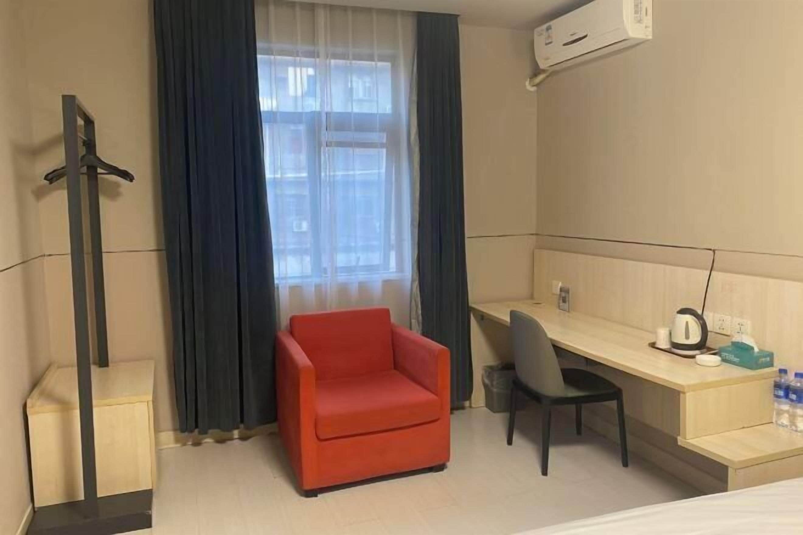 Habitación de hotel con cama, escritorio, silla, sillón rojo, ventana con cortinas y aire acondicionado de pared.