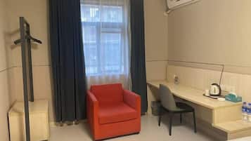 Habitación de hotel con cama, escritorio, silla, sillón rojo, ventana con cortinas y aire acondicionado de pared.