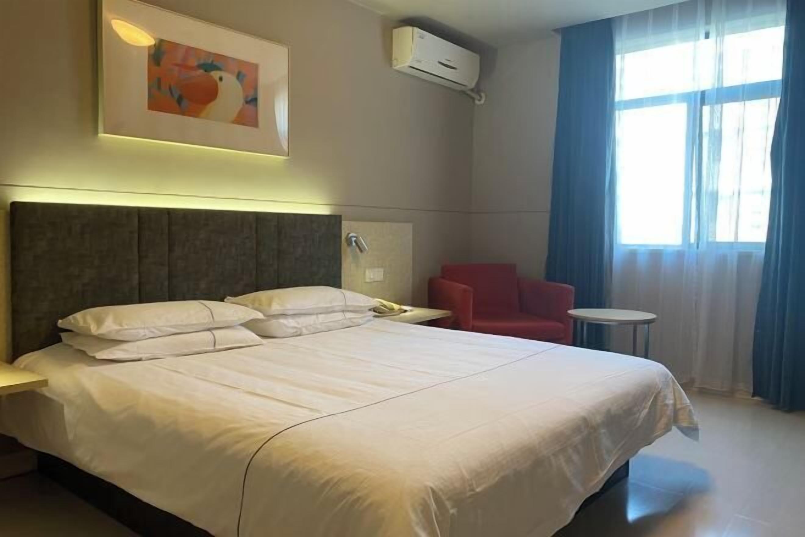 A hotel room with a large bed, a red chair, a small round table, and a window with curtains.