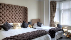 A hotel room with a large tufted headboard, two beds with dark and light bedding, a chair, and a window with vertical blinds. - Kildonan Guesthouse (Aberdeen)