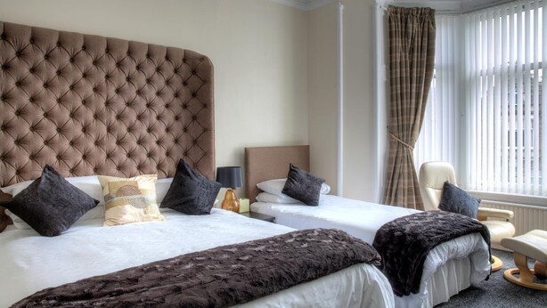 A hotel room with a large tufted headboard, two beds with dark and light bedding, a chair, and a window with vertical blinds. - Kildonan Guesthouse (Aberdeen)