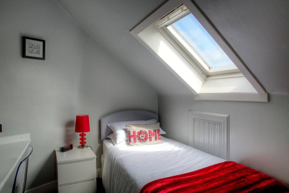 Une chambre avec un puits de lumière, un lit recouvert d’une couverture rouge, une table de chevet avec une lampe et une bande dessinée encadrée accrochée au mur.