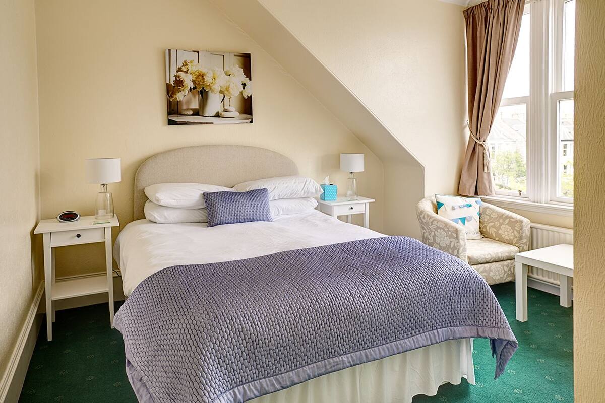 Une chambre à coucher avec un lit, deux tables de chevet, une chaise, une fenêtre avec des rideaux et un tableau au mur.