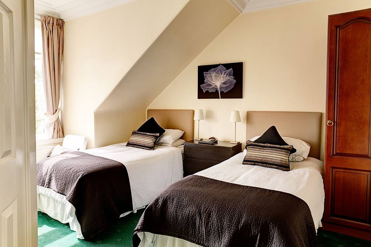 Une chambre d’hôtel avec deux lits, un tableau au mur, une porte en bois et une fenêtre avec des rideaux.