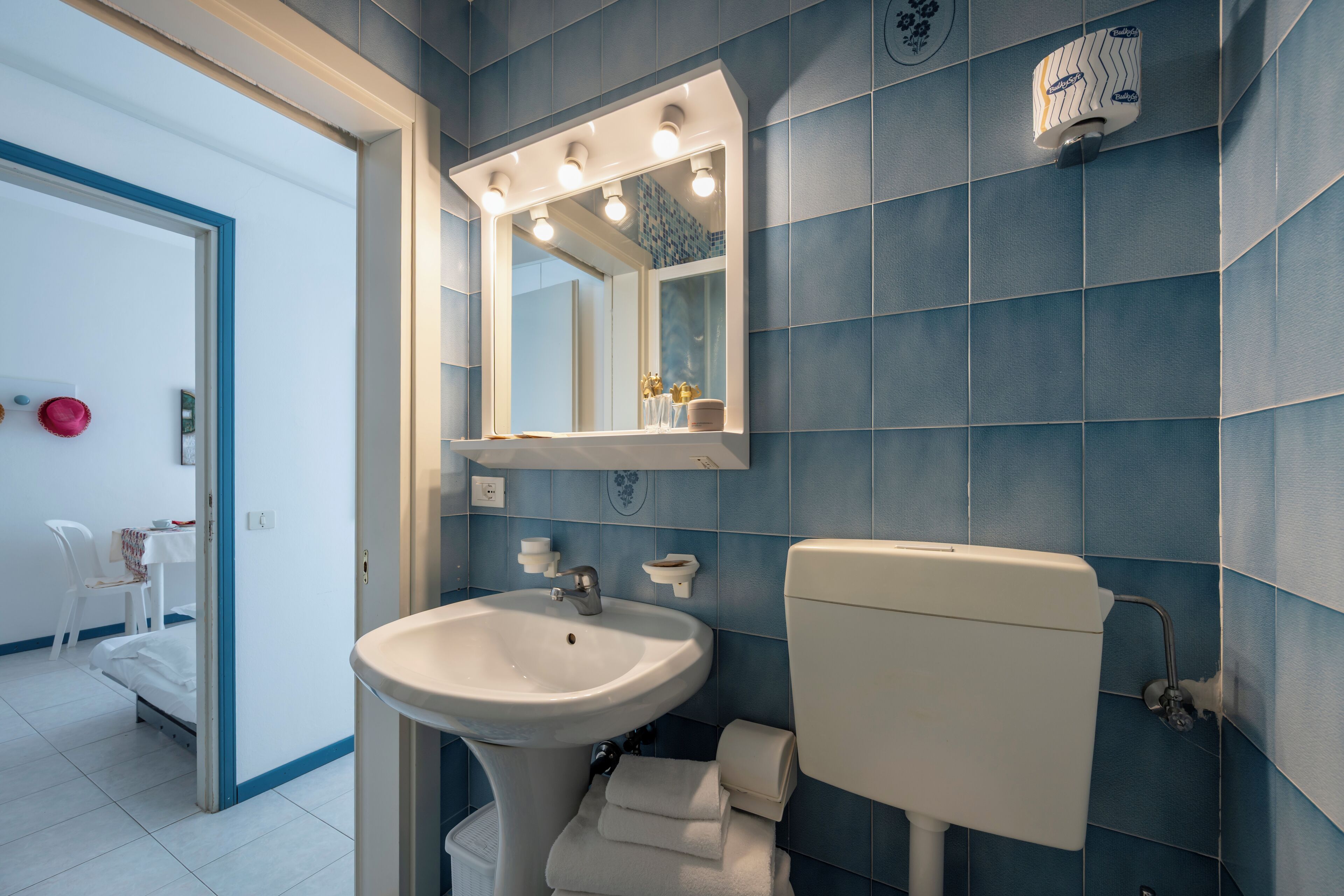 A bathroom with a white sink, mirror, and toilet. There is a blue tiled wall and a white door leading to a bedroom.