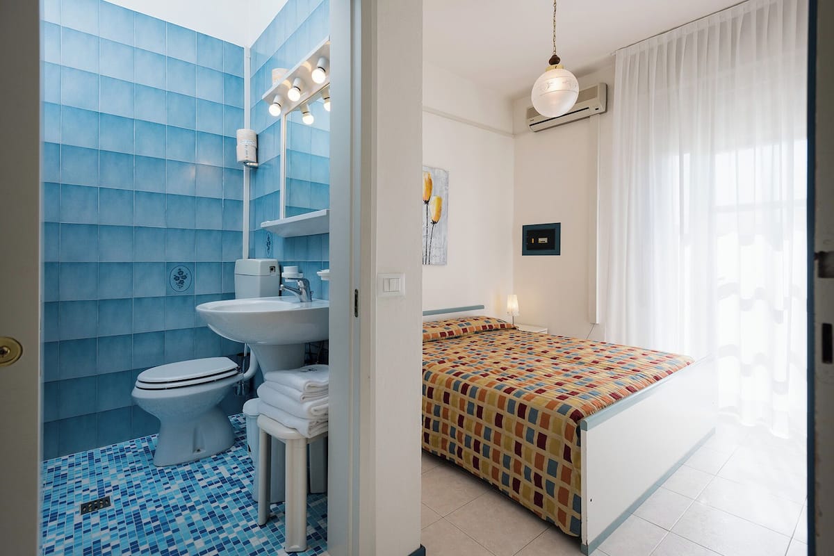 Een badkamer met toilet, wastafel en douche. Er staat een bed met een kleurrijk dekbed en een nachtkastje met een lamp.