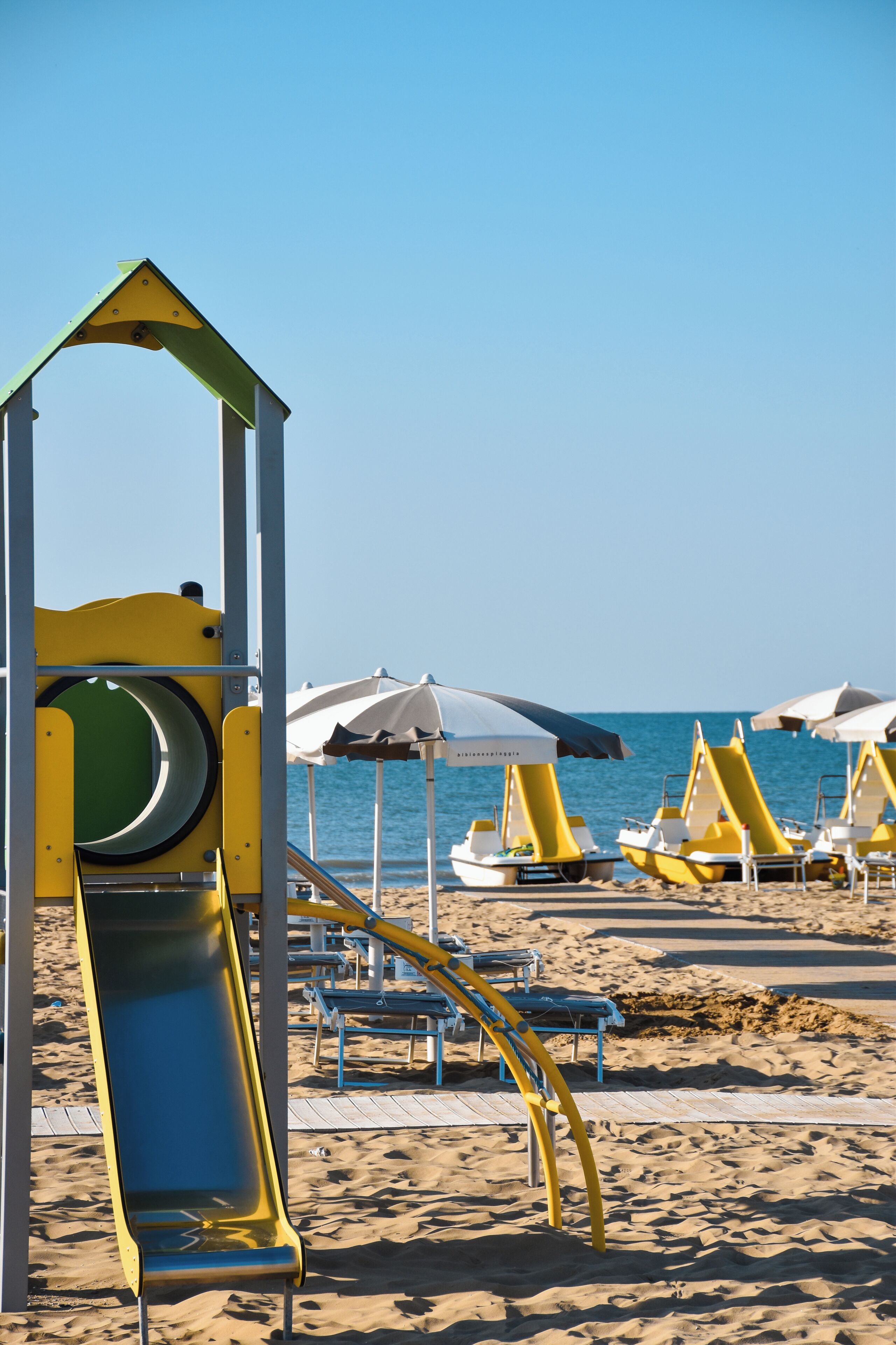 Une plage avec des transats jaunes et blancs, un toboggan jaune et vert, et des parasols blancs.